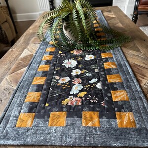 Puede incluir: Un camino de mesa floral blanco y negro con un borde amarillo y negro. El camino de mesa est&aacute; sobre una mesa de madera con una planta en una maceta.