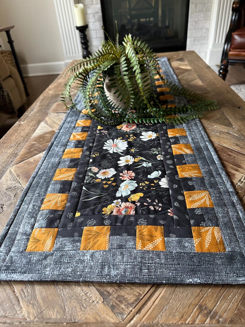 Puede incluir: Un camino de mesa floral blanco y negro con un borde amarillo y negro. El camino de mesa est&aacute; sobre una mesa de madera con una planta en una maceta.