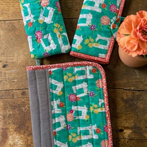 May include: Three quilted fabric items in a teal color featuring a pattern of white sewing machines, yellow citrus fruits, and pink flowers. The items include a zippered pouch, a folding wallet, and a larger rectangular organizer case.