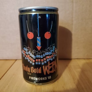 May include: A black aluminum can of Muddy Gold Beer with a red, white, and blue design. The can has a cherry and firework graphic and the text "Fireworks '85".