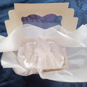 May include: A white eyelet dress is nestled in a box with tissue paper. The box is open, revealing a glimpse of purple fabric. The dress has a pleated skirt and eyelet detailing.