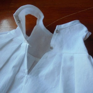 May include: Two white baby doll dresses with pleated skirts and short sleeves. The dresses have snap closures at the neck and are made of a lightweight fabric. The dresses are likely for a baby or toddler.