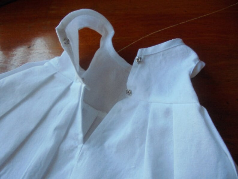 May include: Two white baby doll dresses with pleated skirts and short sleeves. The dresses have snap closures at the neck and are made of a lightweight fabric. The dresses are likely for a baby or toddler.