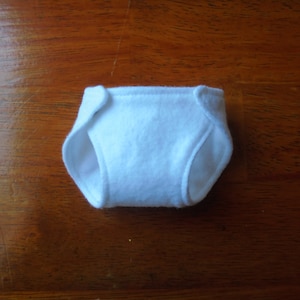 May include: A small, white cloth diaper with curved edges and tabs. The diaper is made of a soft, absorbent material, and is designed for a baby doll or toy. The diaper is shown on a wooden surface.