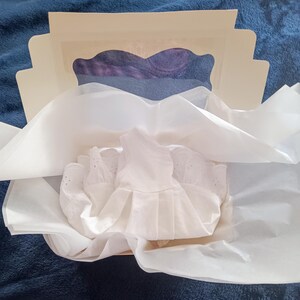 May include: A white eyelet dress is nestled in a box with tissue paper. The box is open, revealing a glimpse of purple fabric. The dress has a pleated skirt and delicate details.