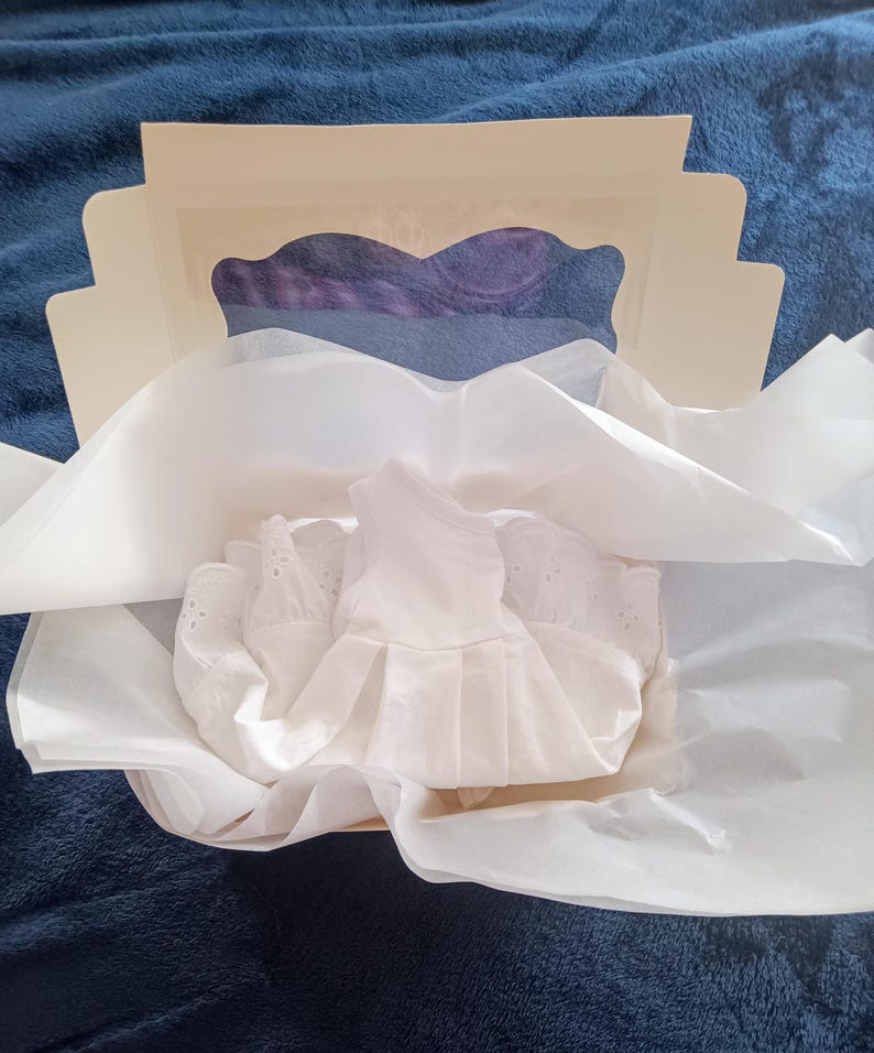 May include: A white eyelet dress is nestled in a box with tissue paper. The box is open, revealing a glimpse of purple fabric. The dress has a pleated skirt and delicate details.