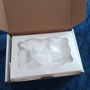 May include: An open cardboard box with a white insert. The insert has a decorative cutout and is filled with white tissue paper. The box is on a blue surface.