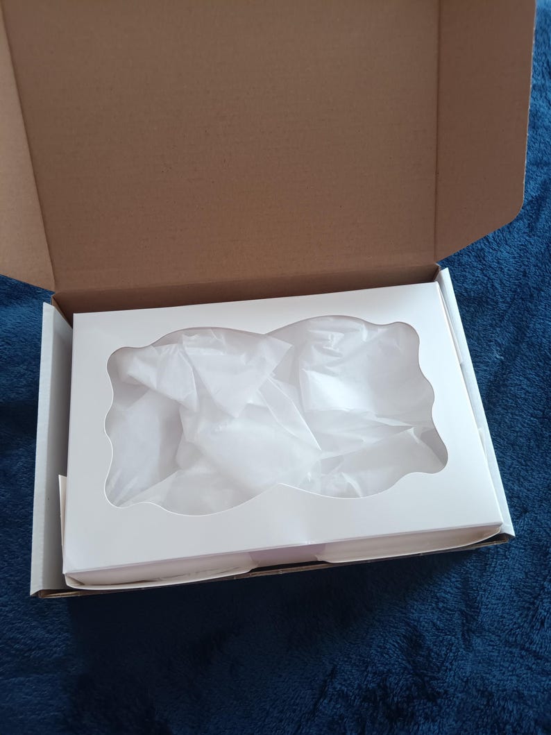 May include: An open cardboard box with a white insert. The insert has a decorative cutout and is filled with white tissue paper. The box is on a blue surface.