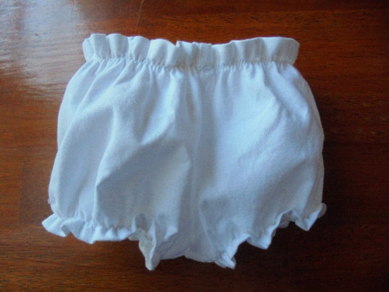 May include: White baby bloomers with a ruffled elastic waistband and leg openings. The bloomers are made of a soft, lightweight fabric, perfect for warm weather. The garment is designed for comfort and ease of movement.
