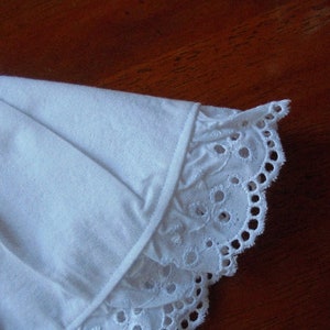 May include: Close-up of a white fabric with a detailed eyelet lace trim. The fabric appears to be cotton or a similar material, with a delicate, scalloped edge. The trim features small, circular cutouts.