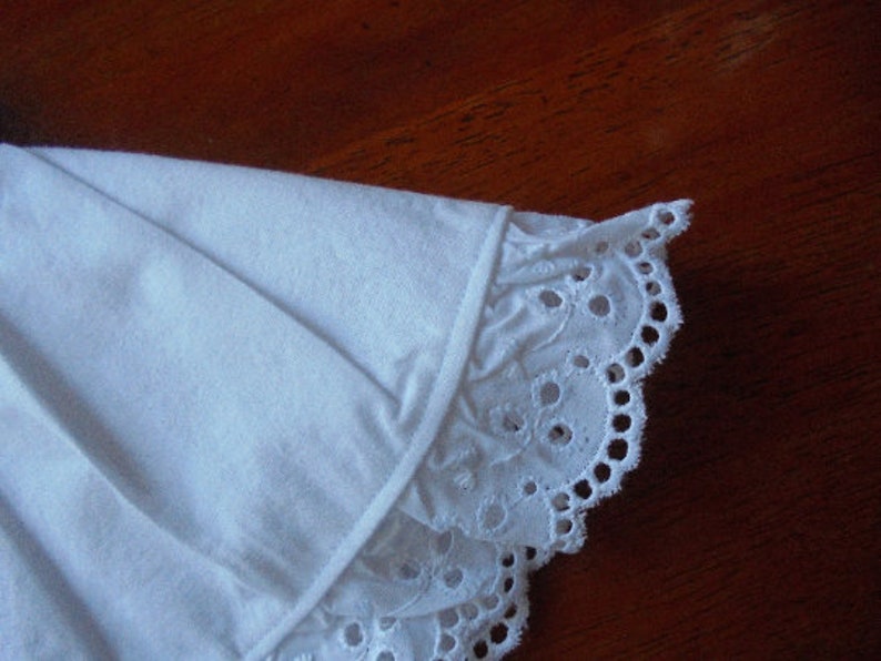 May include: Close-up of a white fabric with a detailed eyelet lace trim. The fabric appears to be cotton or a similar material, with a delicate, scalloped edge. The trim features small, circular cutouts.