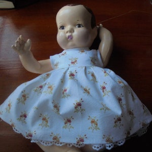 May include: A vintage baby doll with a painted face and brown hair, wearing a white dress with a floral pattern in shades of red and yellow. The dress has a lace trim around the hem.
