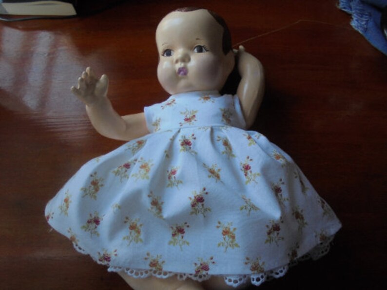 May include: A vintage baby doll with a painted face and brown hair, wearing a white dress with a floral pattern in shades of red and yellow. The dress has a lace trim around the hem.