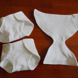Puede incluir: Tres componentes de pañales de tela blancos se muestran sobre una superficie marrón. Dos son pañales plegados con cierres de velcro, y el tercero es un inserto contorneado. Los artículos están hechos de un material suave y absorbente.