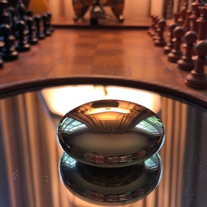 Puede incluir: Un objeto metálico ovalado y pulido que refleja la habitación, incluyendo un tablero de ajedrez y piezas de ajedrez. El objeto está sobre una superficie espejada, con un marco de madera. Las piezas de ajedrez son negras y marrones.