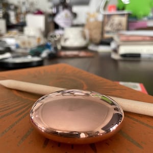 Puede incluir: Un objeto ovalado brillante de color oro rosa descansa sobre una superficie texturizada de color naranja-marrón. Un palo de madera de color claro está colocado diagonalmente sobre la superficie. El fondo está borroso, lo que sugiere un escritorio o espacio de trabajo.