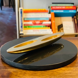 May include: A modern, metallic gold-colored desk organizer with two elongated compartments, resting on a black, circular base. The organizer has a sleek, minimalist design, reflecting light. Bookshelves are visible in the background.