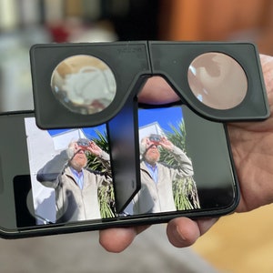 Könnte beinhalten: Schwarzes VR-Headset mit kreisförmigen Linsen, das an einem Smartphone befestigt ist, das ein Bild anzeigt. Das Headset wird in einer Hand gehalten, und die Linsen sind vor dem Bildschirm des Telefons positioniert. Das Bild auf dem Bildschirm zeigt eine Person.