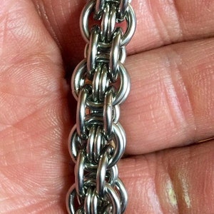 May include: A silver chain bracelet with a woven design. The bracelet is made of small, interlocking metal rings.