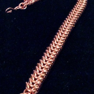 Op de afbeelding: Een koperen kettingbaard armband met een geweven ontwerp. De armband is gemaakt van kleine, in elkaar grijpende metalen ringen.