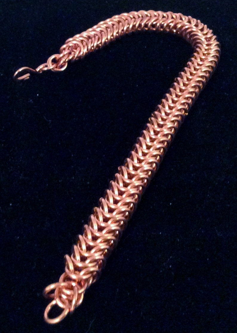 Op de afbeelding: Een koperen kettingbaard armband met een geweven ontwerp. De armband is gemaakt van kleine, in elkaar grijpende metalen ringen.