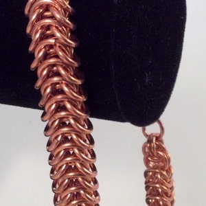 Op de afbeelding: Een koperen kettingbaard armband met een geweven patroon. De armband is op een zwarte fluwelen displaystandaard.