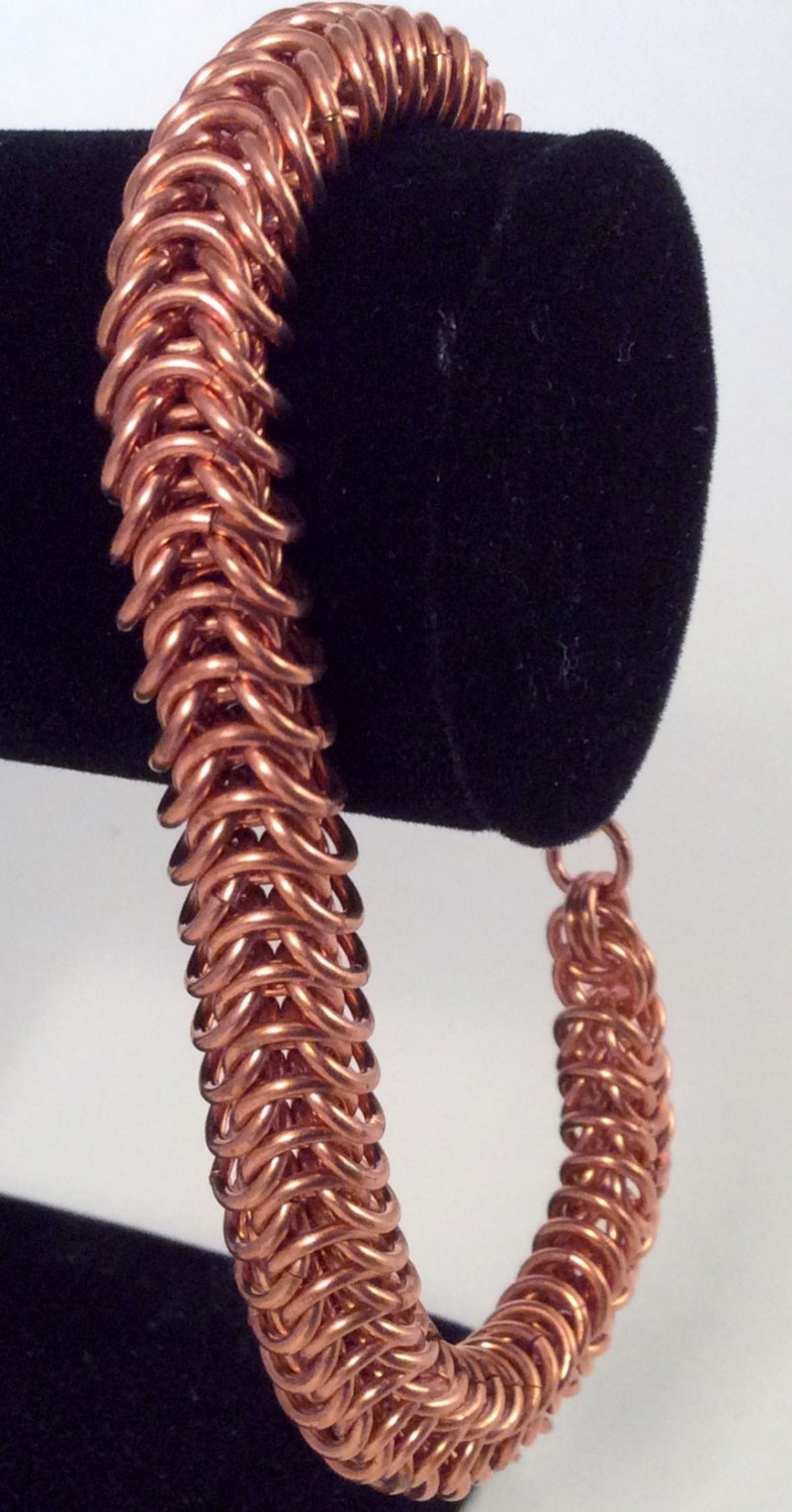 Op de afbeelding: Een koperen kettingbaard armband met een geweven patroon. De armband is op een zwarte fluwelen displaystandaard.