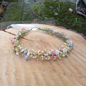 以下が含まれることがあります： 白、ピンク、青、黄色の乾燥花でできた繊細な花冠。ワイヤーベースで作られており、白いリボンで結ばれています。