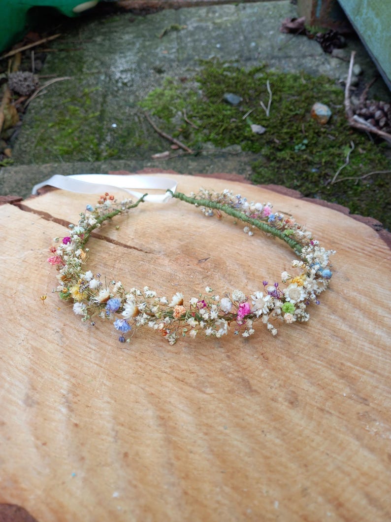 以下が含まれることがあります： 白、ピンク、青、黄色の乾燥花でできた繊細な花冠。ワイヤーベースで作られており、白いリボンで結ばれています。