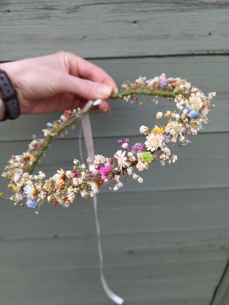 以下が含まれることがあります： 白、ピンク、青、緑の乾燥花でできた繊細な花冠。白いリボンで結ばれています。