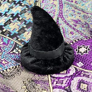 May include: A small, black plush witch hat with a wide brim and a black band. The hat is set against a patterned background with shades of purple, blue, and gold. A festive accessory for Halloween.
