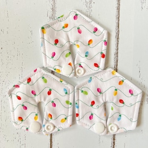 May include: Three white fabric nursing pads with a festive Christmas lights pattern. Each pad features a U-shaped cutout and a white snap closure. The pads are decorated with colorful lights on a white background.