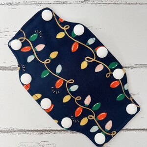 May include: A navy blue fabric item with a pattern of colorful Christmas lights and white snap closures. The lights are red, green, yellow, and pink, with gold-colored string. The item is likely a reusable cloth product.