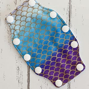 May include: A blue and purple fabric with a mermaid scale pattern and white snap buttons around the edges.