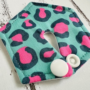 Könnte beinhalten: Ein türkisfarbenes Stoffteil mit einem rosa und dunkel türkisfarbenen Leopardenmuster. Der Artikel hat einen U-förmigen Ausschnitt und zwei weiße Druckknöpfe. Der Artikel liegt auf einer weißen Holzoberfläche.