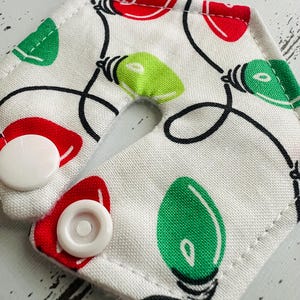May include: A white fabric item with a Christmas lights pattern in red and green, connected by black lines. The item has a hexagonal shape with two white snap closures. The design is festive and holiday-themed.