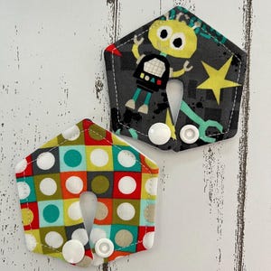 May include: Two fabric neck pillows. One is patterned with a robot and stars on a gray background. The other has a colorful geometric pattern of squares and circles. Both have white snaps.