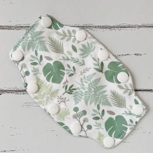 May include: A white cloth baby bib with a botanical print of green leaves and ferns. The bib has a curved shape and is secured with seven white snaps.