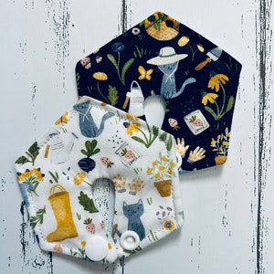 May include: Two pentagon-shaped nursing pads with a cat and garden-themed print. One pad has a white background with yellow flowers, a yellow boot, and a gray cat. The other pad has a navy background with a cat wearing a hat.