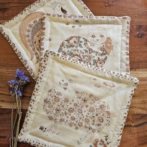 May include: Three square fabric coasters in cream with brown floral illustrations. One features a sheep, another a cat with a bow, and the third a bird. Each has a decorative border with small heart and animal motifs, and a quilted texture.