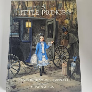 May include: A hardcover book titled A Little Princess by Frances Hodgson Burnett, illustrated by Graham Rust. The cover features a young person in a blue coat standing by a horse-drawn carriage, with a person carrying luggage nearby at night.