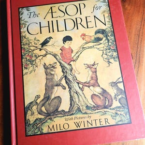 May include: A hardcover book titled The AESOP for CHILDREN with a red cover and a central illustration featuring a child sitting in a tree surrounded by animals including a crow, rooster, fox, wolf, tortoise, and rabbit, with art by Milo Winter.