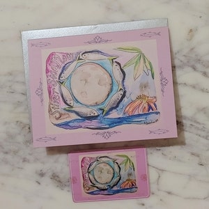 May include: A pink card with a silver border features a watercolor illustration of a full moon surrounded by silver fish. The text "Silver Shagm" is written in a cursive font on the left side of the card.