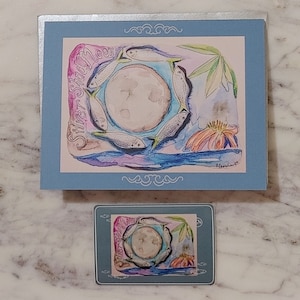 May include: A watercolor painting of a full moon with a circle of silver fish swimming around it. The painting is on a light blue background with a silver border. The text "Silver Slam Moon" is written in a cursive font on the left side of the painting.