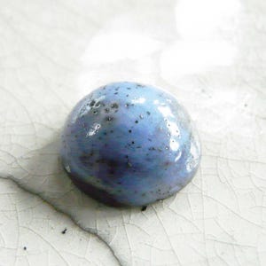 Könnte beinhalten: Eine kleine, runde, blaue Keramikperle mit einem gesprenkelten Muster. Die Perle hat eine glänzende Oberfläche und liegt auf einer weißen, rissigen Oberfläche. Die Perle hat einen Durchmesser von etwa 2 cm.