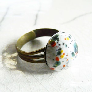 May include: A bronze-toned ring with a speckled, teardrop-shaped ceramic stone. The stone is primarily white with colorful speckles of red, yellow, green, and blue. The ring band has a triple-band design.