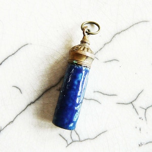 May include: A cylindrical pendant featuring a deep blue, textured ceramic body capped with a metallic, brass-toned top. The top includes a small, circular loop for attachment, showcasing a weathered, aged finish on the metal component.