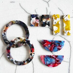 May include: An assortment of colorful acrylic jewelry components. The pieces include two circular rings, two pairs of rectangular earrings, and two half-circle pendants. The colors are a mix of blue, red, yellow, black, and white.