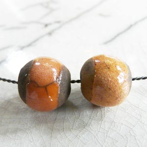 Puede incluir: Dos cuentas de cerámica hechas a mano. Cada cuenta es una mezcla de colores marrón, naranja y beige. Las cuentas están ensartadas en un alambre fino y oscuro. Las cuentas tienen un acabado brillante y un aspecto texturizado.