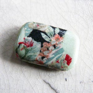 Può includere: Piccolo oggetto rettangolare decorativo con motivo floreale. L'oggetto ha uno sfondo verde chiaro con un disegno di fiori e foglie rosse, nere e arancioni. L'oggetto è su una superficie bianca strutturata.